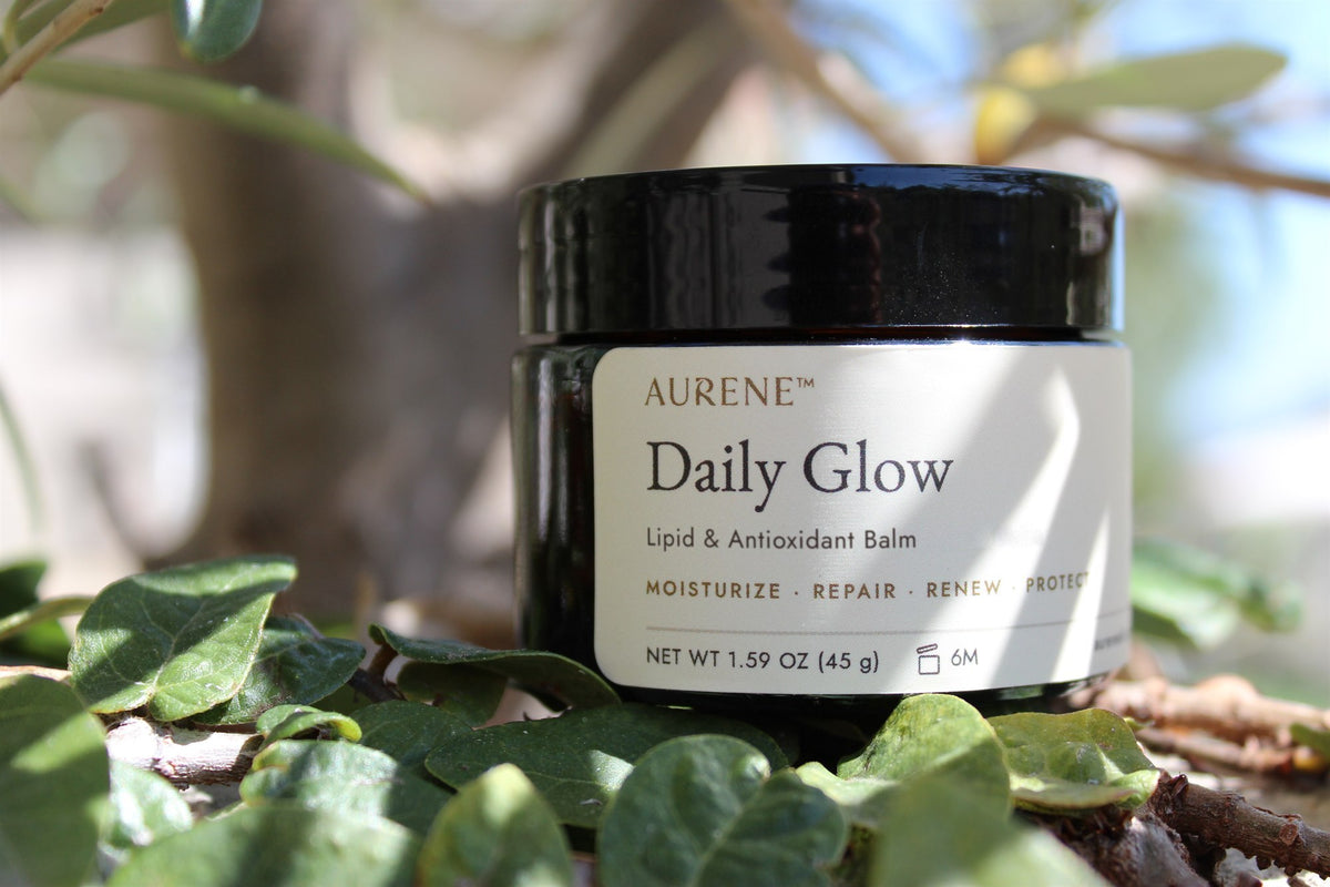 A closed amber glass jar of Aurene Daily Glow balm resting on green leaves in dappled sunlight.